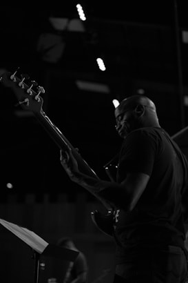 A musician is playing a bass guitar on stage under dim lighting. The focus is on the instrument and the upper body of the musician. The background is dark, with spotlights illuminating partially visible stage equipment.