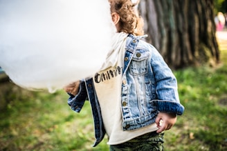 A child with curly hair is wearing a denim jacket over a light-colored shirt that has writing on it. They are holding a large white cotton candy in front of their face. The background is grassy with a tree trunk visible, suggesting an outdoor setting.
