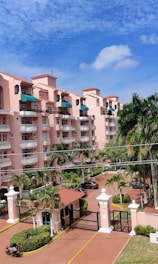 A multi-story residential building with pink stucco walls and terracotta-style roofs. The building features several balconies, many with green awnings. In the foreground, there are tall palm trees and a gated entrance with a guardhouse that includes a small covered structure. The sky above is mostly clear with a few scattered clouds.