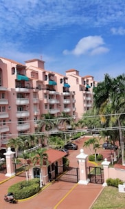 A multi-story residential building with pink stucco walls and terracotta-style roofs. The building features several balconies, many with green awnings. In the foreground, there are tall palm trees and a gated entrance with a guardhouse that includes a small covered structure. The sky above is mostly clear with a few scattered clouds.