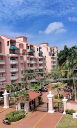 A multi-story residential building with pink stucco walls and terracotta-style roofs. The building features several balconies, many with green awnings. In the foreground, there are tall palm trees and a gated entrance with a guardhouse that includes a small covered structure. The sky above is mostly clear with a few scattered clouds.