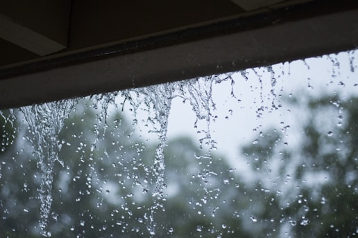 Rainwater cascades down from the edge of a rooftop or overhang, with a blurred background of trees and overcast skies.