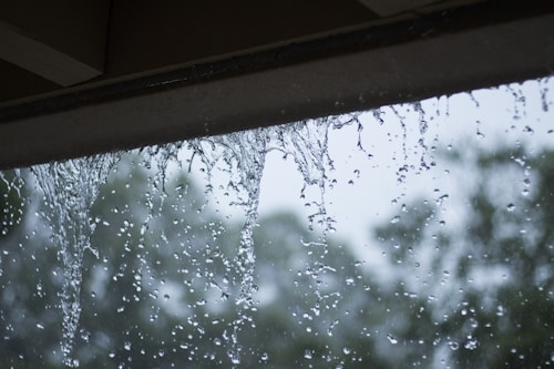 Rainwater cascades down from the edge of a rooftop or overhang, with a blurred background of trees and overcast skies.
