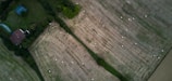 An aerial view of agricultural fields with visible patches of green grass and numerous hay bales scattered across the brownish, harvested areas. A small house with a reddish roof is nestled among trees, surrounded by verdant vegetation.