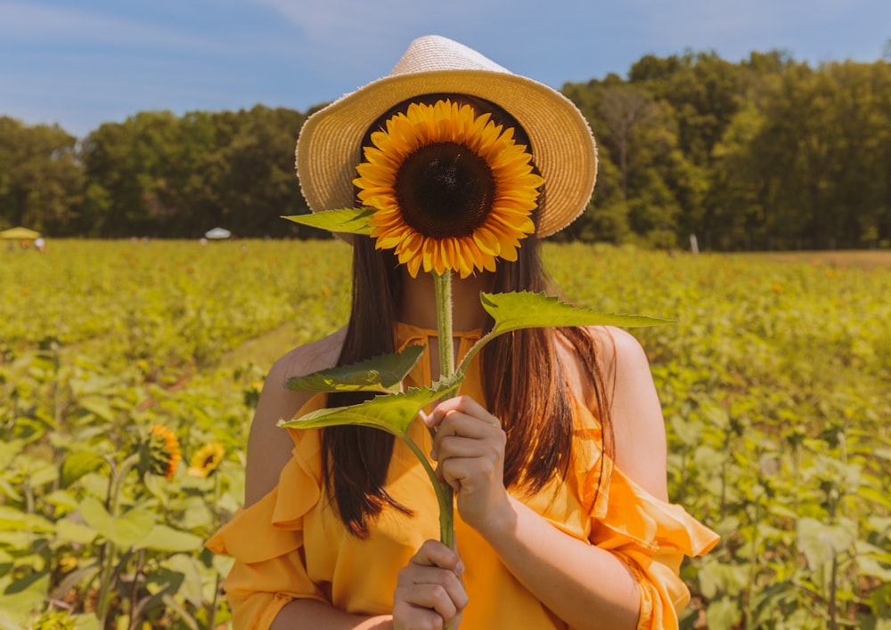 Frau, die tagsüber ihr Gesicht mit Sonnenblume auf dem Feld in der Nähe von Bäumen bedeckt