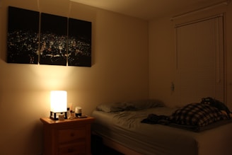 A dimly lit bedroom features a single bed with grey bedding and a plaid blanket. A wooden nightstand next to the bed holds a cube-shaped lamp emitting a warm glow, along with some small items. Three-part wall art depicting a cityscape at night is mounted above the bed. A window with closed blinds is situated on the right side of the room.