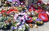 A variety of vibrant floral wreaths and arrangements are lying on the ground, accompanied by ribbons with the words 'Lest We Forget.' Some wreaths feature bold red poppies, while others include mixed flowers such as roses and proteas. There is a photograph and a card referencing World War Two Service among the flowers.