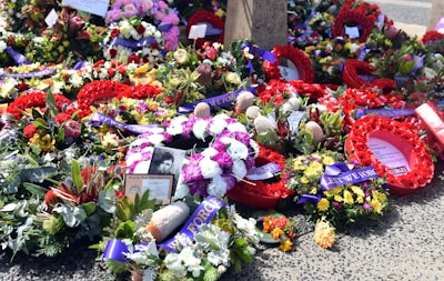 A variety of vibrant floral wreaths and arrangements are lying on the ground, accompanied by ribbons with the words 'Lest We Forget.' Some wreaths feature bold red poppies, while others include mixed flowers such as roses and proteas. There is a photograph and a card referencing World War Two Service among the flowers.