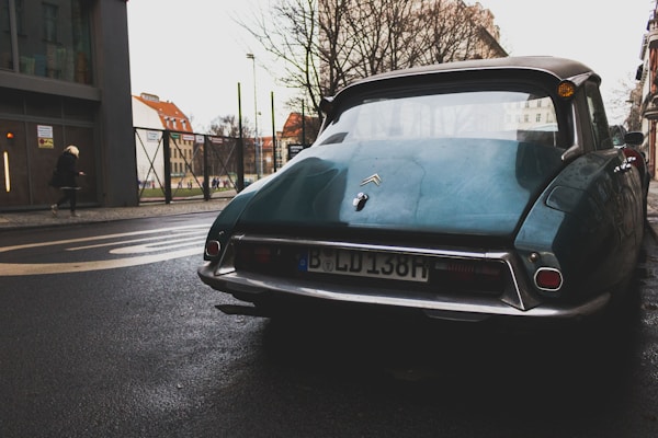 A vintage Citroën car parked on a street with wet pavement. The setting includes urban elements such as buildings, a fence, and a passerby with a modern attire. Bare trees suggest a winter or early spring atmosphere.