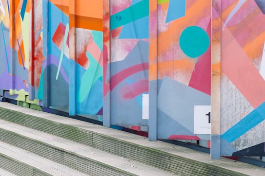 A vibrant wall covered in abstract graffiti art with geometric shapes and patterns in various colors, including orange, blue, pink, and red. The design includes a large turquoise circle with drips and is set against metal panels. The scene is framed by a set of wooden steps leading up to the artwork.