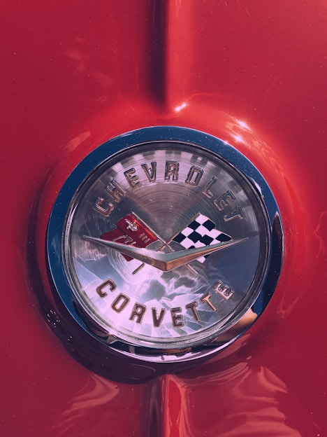 The image features a close-up of a classic Chevrolet Corvette emblem set against a glossy red surface. The emblem displays two crossed flags: one checkered and the other with a Chevrolet logo. Chrome detailing surrounds the emblem, adding a vintage and polished look.