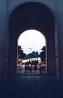 A large archway frames a view of a spherical structure with the word 'UNIVERSAL' lit up on its surface. People can be seen walking around the area, and the scene is set against a backdrop of palm trees and twilight sky.