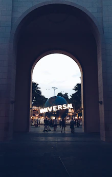 A large archway frames a view of a spherical structure with the word 'UNIVERSAL' lit up on its surface. People can be seen walking around the area, and the scene is set against a backdrop of palm trees and twilight sky.