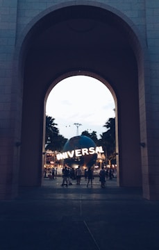 A large archway frames a view of a spherical structure with the word 'UNIVERSAL' lit up on its surface. People can be seen walking around the area, and the scene is set against a backdrop of palm trees and twilight sky.