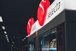 A vending machine with a red circular logo displaying the brand name 'selecta' illuminated at the top. Various snacks are visible inside the machine, with dim lighting in the surrounding environment.