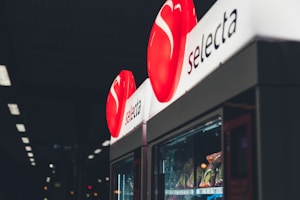 A vending machine with a red circular logo displaying the brand name 'selecta' illuminated at the top. Various snacks are visible inside the machine, with dim lighting in the surrounding environment.