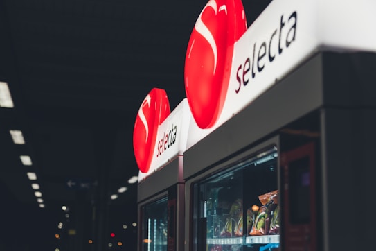 A vending machine with a red circular logo displaying the brand name 'selecta' illuminated at the top. Various snacks are visible inside the machine, with dim lighting in the surrounding environment.