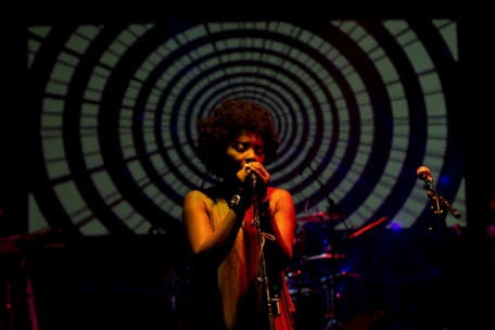A singer is performing on stage with a microphone, illuminated by warm light. The background features a large circular, hypnotic pattern with concentric rings. The stage is dimly lit, enhancing the focus on the performer, whose face is partially obscured by shadows.