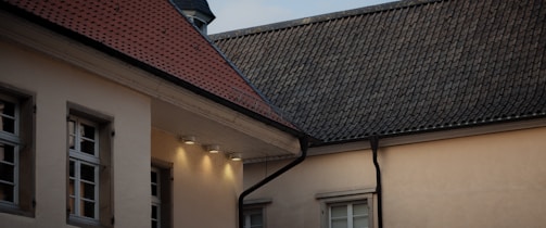 A section of a building featuring traditional sloped roofs with tiles. The walls are light beige, contrasted by the dark roofing materials. The windows are symmetrically placed in wooden frames, and lighting fixtures are installed below the roof overhang, illuminating the building wall with a warm glow.