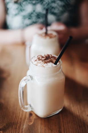 Two creamy beverages topped with chocolate sprinkles served in mason jars with handles and black straws. The drinks are placed on a wooden surface, with a blurred background showing a person holding one of the jars.