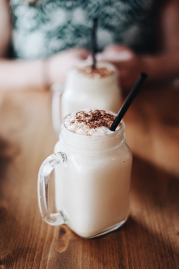 Two creamy beverages topped with chocolate sprinkles served in mason jars with handles and black straws. The drinks are placed on a wooden surface, with a blurred background showing a person holding one of the jars.