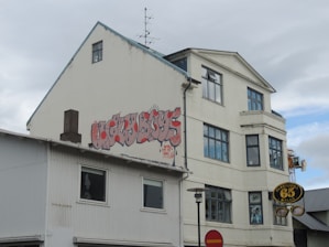 A three-story building with graffiti on the side reading 'Ugly Boys' in stylized red letters. The building is a mix of white and gray tones. There are multiple large windows, and a small antenna is visible on the roof. A circular sign hangs in front of the building with the text 'Oleraugna 65 Sala'. The sky is overcast with light clouds.