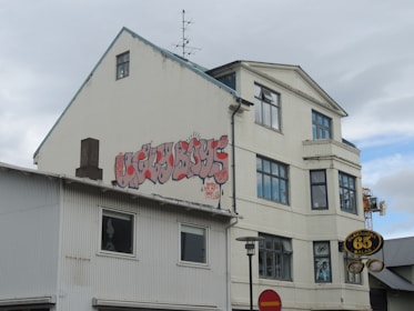 A three-story building with graffiti on the side reading 'Ugly Boys' in stylized red letters. The building is a mix of white and gray tones. There are multiple large windows, and a small antenna is visible on the roof. A circular sign hangs in front of the building with the text 'Oleraugna 65 Sala'. The sky is overcast with light clouds.