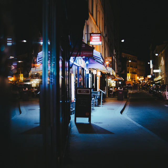 Cars parked in front of lit shops and bars on a Paris street at night
