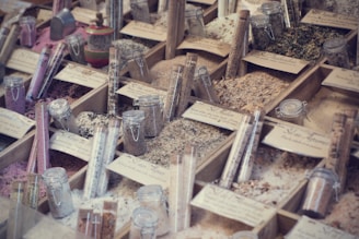 A display of various types of salts and spices organized in wooden boxes, with each section labeled. Glass jars and test tubes filled with different colored contents are placed among the boxes, showcasing a variety of textures and colors, from pink to green to white.
