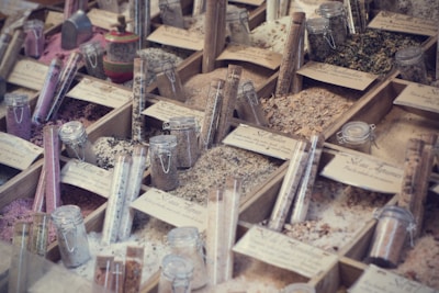 A display of various types of salts and spices organized in wooden boxes, with each section labeled. Glass jars and test tubes filled with different colored contents are placed among the boxes, showcasing a variety of textures and colors, from pink to green to white.