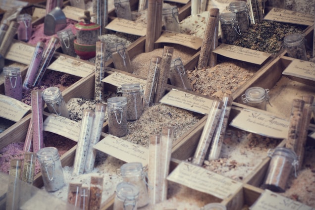 A display of various types of salts and spices organized in wooden boxes, with each section labeled. Glass jars and test tubes filled with different colored contents are placed among the boxes, showcasing a variety of textures and colors, from pink to green to white.