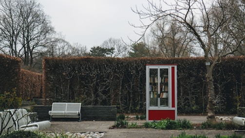 A small outdoor library with a red and white frame stands in a well-maintained garden area. Leafless trees and trimmed hedges form the background, while white benches are arranged around the space. The scene has an overcast sky, and small plants and rocks decorate the ground.