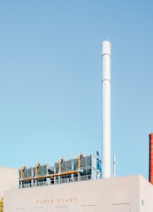 A large industrial building marked with the words 'Power Plant' features a tall, cylindrical chimney stack. The building's roof is equipped with metal fencing and industrial piping. The sky is a clear, pale blue.