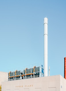 A large industrial building marked with the words 'Power Plant' features a tall, cylindrical chimney stack. The building's roof is equipped with metal fencing and industrial piping. The sky is a clear, pale blue.