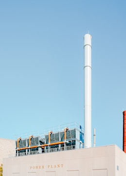 A large industrial building marked with the words 'Power Plant' features a tall, cylindrical chimney stack. The building's roof is equipped with metal fencing and industrial piping. The sky is a clear, pale blue.