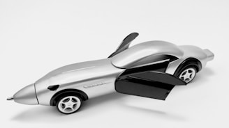 A sleek, futuristic toy car with a metallic finish. The car has two open gull-wing doors and four small wheels. The design is streamlined with a pointed front and rounded back, resembling a fusion between a car and an airplane.