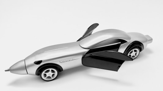 A sleek, futuristic toy car with a metallic finish. The car has two open gull-wing doors and four small wheels. The design is streamlined with a pointed front and rounded back, resembling a fusion between a car and an airplane.