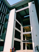 A modern architectural structure with multiple levels and balconies, characterized by clean lines and a mix of light and shadow. The building facade features large rectangular windows and smooth surfaces with a muted color palette. A pair of recycling bins is visible at the bottom left corner, adding a splash of color.