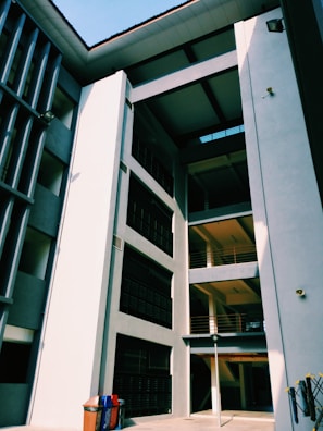 A modern architectural structure with multiple levels and balconies, characterized by clean lines and a mix of light and shadow. The building facade features large rectangular windows and smooth surfaces with a muted color palette. A pair of recycling bins is visible at the bottom left corner, adding a splash of color.