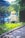 An elderly person with short blond hair is sitting by a canal or river, surrounded by greenery and trees. The setting appears to be a park or a garden with a building in the background. Sunlight filters through the trees, creating a serene and peaceful atmosphere.