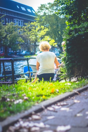 An elderly person with short blond hair is sitting by a canal or river, surrounded by greenery and trees. The setting appears to be a park or a garden with a building in the background. Sunlight filters through the trees, creating a serene and peaceful atmosphere.