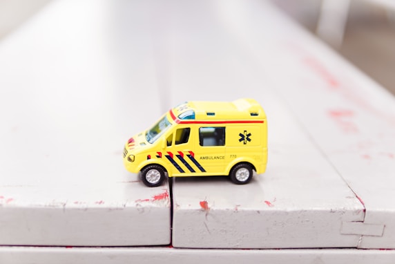 A small toy ambulance painted in yellow with red and blue stripes, positioned on a white surface 