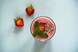 A refreshing strawberry smoothie in a glass garnished with a sliced strawberry, chia seeds, and fresh mint leaves. Two red strawberries are placed on the side, all set on a light blue background.