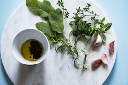 A marble surface features a small bowl of olive oil and balsamic vinegar, surrounded by a variety of fresh herbs including sage, thyme, and oregano. Garlic cloves are arranged artistically beside the bowl. The background is a soft blue color, complementing the fresh and natural theme.