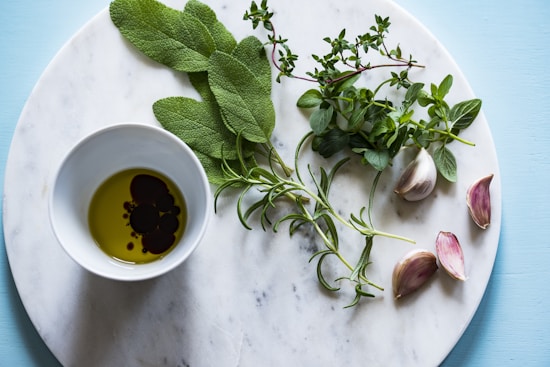 A marble surface features a small bowl of olive oil and balsamic vinegar, surrounded by a variety of fresh herbs including sage, thyme, and oregano. Garlic cloves are arranged artistically beside the bowl. The background is a soft blue color, complementing the fresh and natural theme.