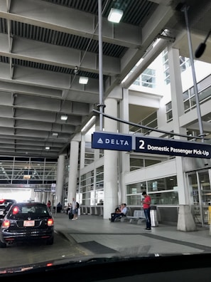 A modern airport terminal area with a focus on the passenger pick-up zone. Several people are waiting, some seated and others standing, under a large overhead sign for Delta Airlines. Cars are lined up at the curbside, and glass windows with vertical columns surround the space.