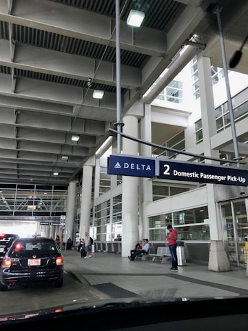 A modern airport terminal area with a focus on the passenger pick-up zone. Several people are waiting, some seated and others standing, under a large overhead sign for Delta Airlines. Cars are lined up at the curbside, and glass windows with vertical columns surround the space.