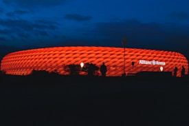 A stadium with a distinctively illuminated facade glows bright red against the backdrop of a dark evening sky. Silhouettes of people and some vegetation are visible in the foreground. The name 'Allianz Arena' is prominently displayed on the stadium.