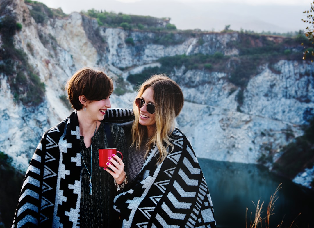 Two women stand closely, wrapped in a blanket, in front of a rocky landscape