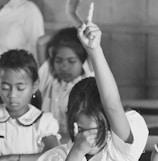 A classroom setting with three children sitting. One child has their hand raised with one finger pointing up, while the other hand covers their face. The other two children have their eyes closed, appearing focused or contemplative.